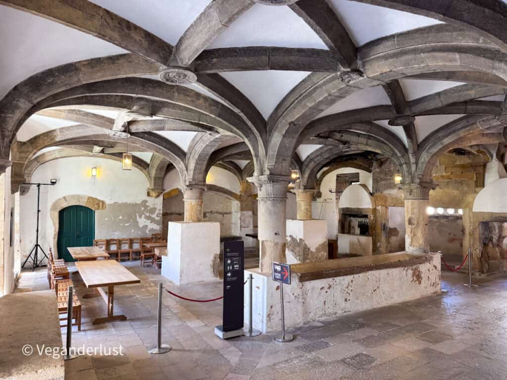 Historic interior room with stone columns and vaulted ceilings supported by arches. Wooden tables and chairs sit beneath the aged plaster walls.