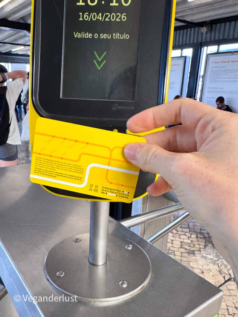 Hand holds a yellow transit card up to a ticket validator machine displaying "10:10 16/04/2026 Valide o seu título" with a green arrow pointing down.