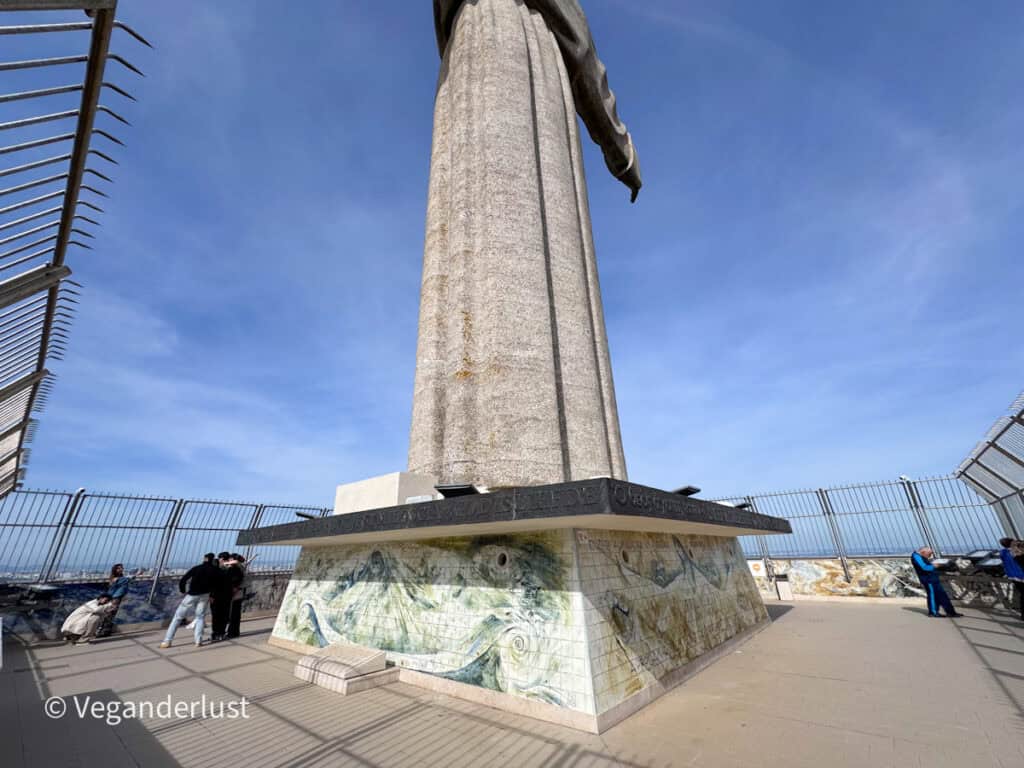 Base of a tall stone statue with outstretched arms stands on a tiled platform surrounded by a fenced viewpoint where small groups of people gather.