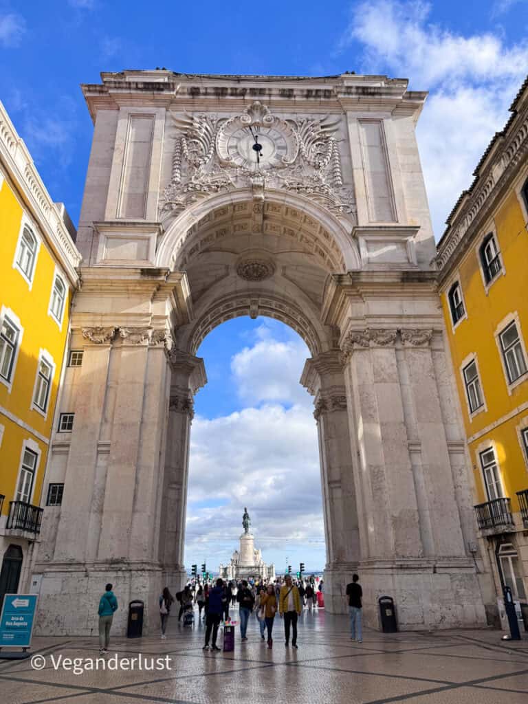 Rua Augusta Arch in Lisbon viewed from below with detailed carvings and clock, framing a busy square with people walking toward the waterfront statue under a blue sky.
