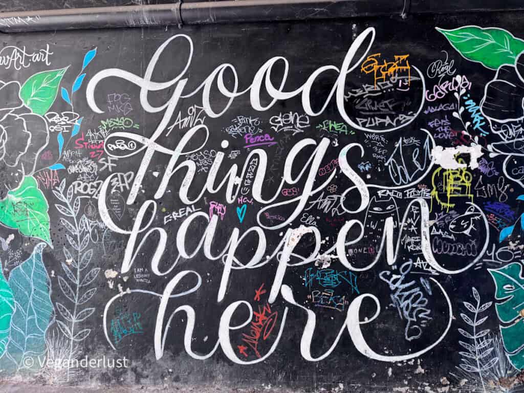 Black wall covered in graffiti and colorful tags features large white script text reading "Good things happen here" surrounded by doodles and leafy illustrations.