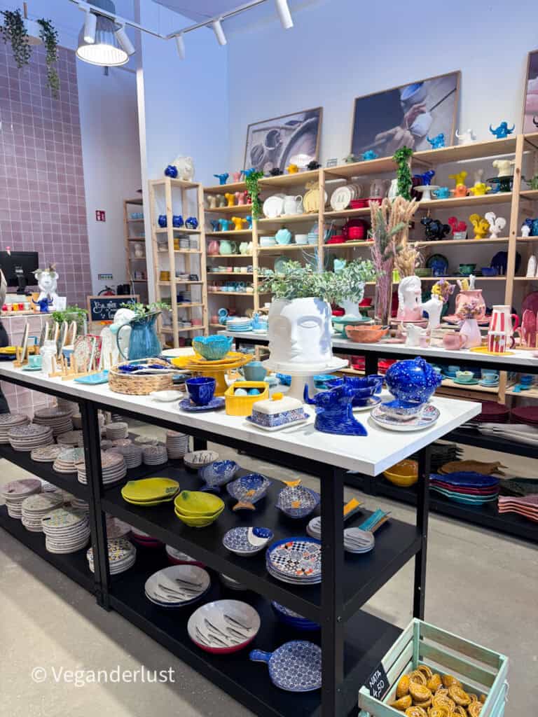 Bright shop interior displaying colorful ceramics including plates, bowls, and decorative pieces arranged on shelves and tables. Various shapes and patterns in blue, yellow, and pastel tones fill the space.