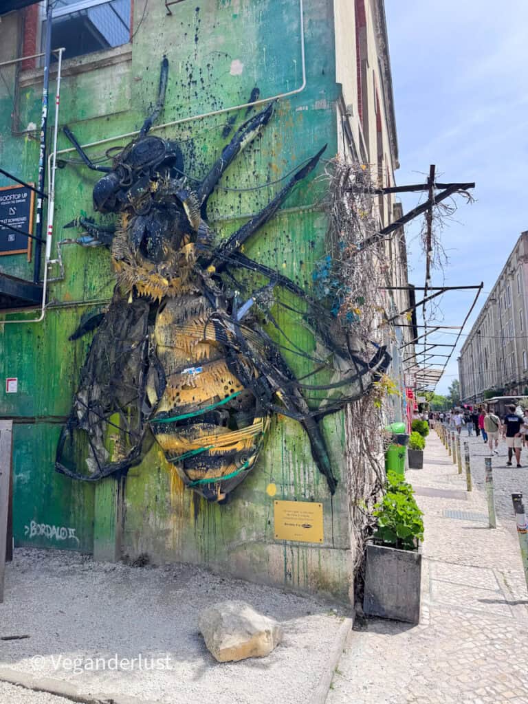 Large street art sculpture of a bee made from metal and found materials mounted on a green wall with paint splatters. A narrow street with pedestrians and buildings extends into the background.