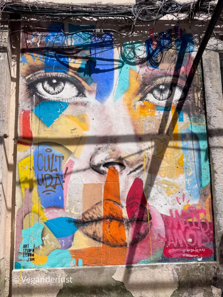 Colorful mural of a woman’s face with detailed eyes and abstract shapes in bright colors covering the wall. Text painted on the mural reads "CUZ? UDA!" along with smaller artist markings.