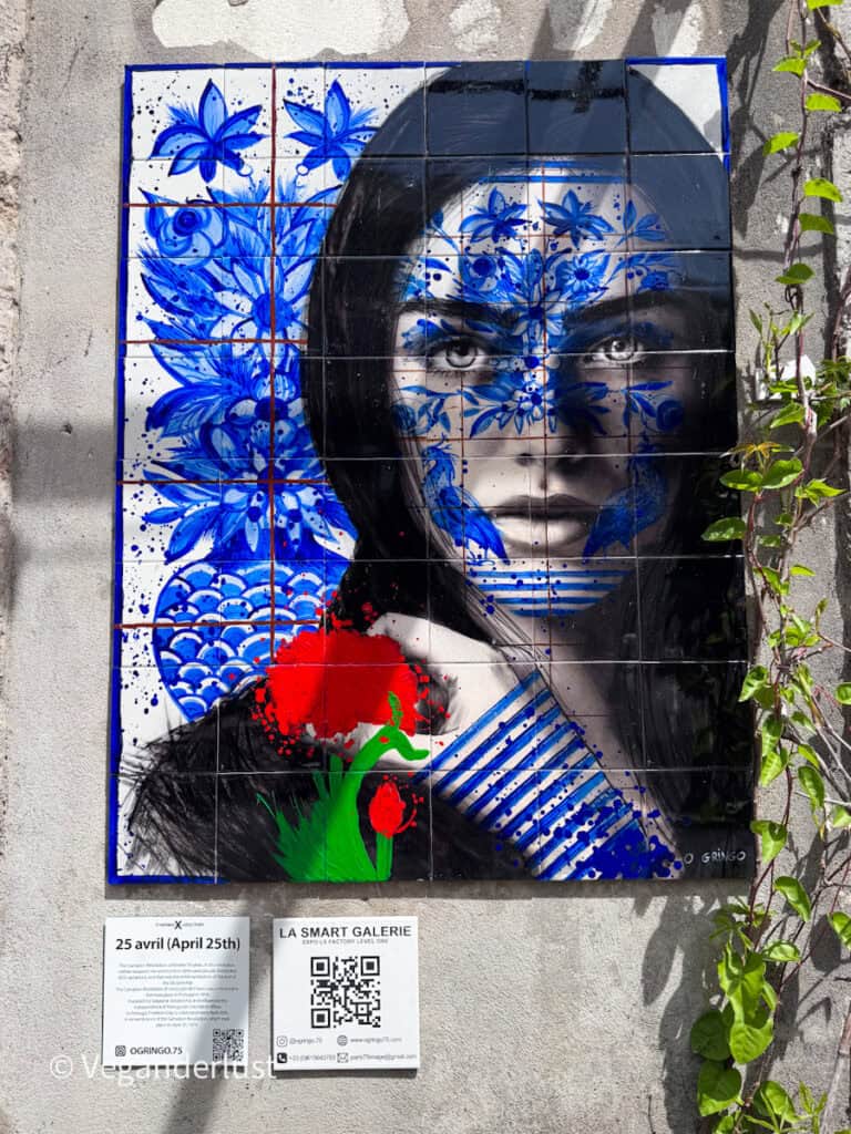 Portrait mural of a woman painted across tiled panels with blue floral patterns covering part of her face and a bright red flower at the bottom. Text below reads "25 abril (April 25th)" and "LA SMART GALERIE" with a QR code and additional small print.
