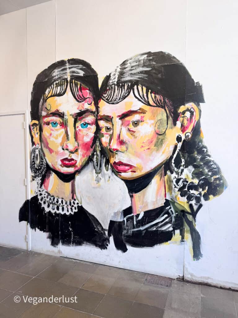 Expressive mural of two women with bold brushstrokes and colorful facial tones painted on a white wall. Their faces lean close together creating an intimate and emotional Lisbon street art scene.
