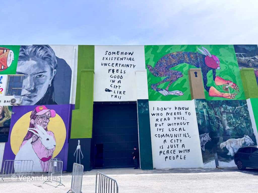 Wide wall covered in mixed street art panels including portraits and surreal figures alongside text that reads "SOMEHOW EXISTENTIAL UNCERTAINTY FEELS GOOD IN A CITY LIKE THIS" and "I DON’T KNOW WHO NEEDS TO READ THIS, BUT WITHOUT ITS LOCAL COMMUNITIES, A CITY IS JUST A PLACE WITH PEOPLE". The colorful composition highlights Lisbon’s diverse street art culture.