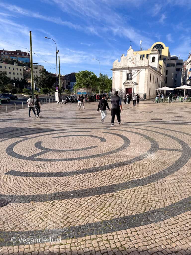 Wide Lisbon square with spiral patterned cobblestone pavement leading toward a small white church and people walking across the open space. The distinctive black and white stone design highlights a classic Lisbon street scene.