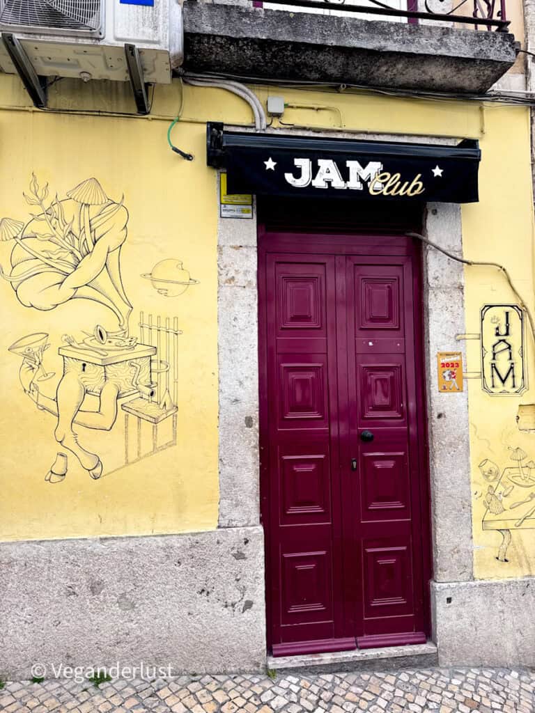 Yellow wall with a red door entrance labeled "JAM Club" surrounded by whimsical line drawings of musical and surreal figures. The worn facade and cobblestone street highlight Lisbon street art culture.