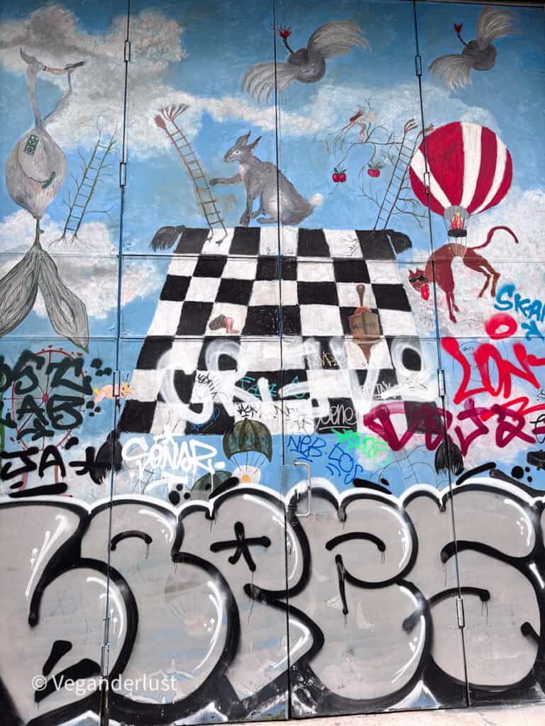 Large mural of surreal animals and figures on a blue sky background above graffiti tags, including a checkerboard platform and a red hot air balloon. Layers of street art and tagging cover the lower portion of the wall.