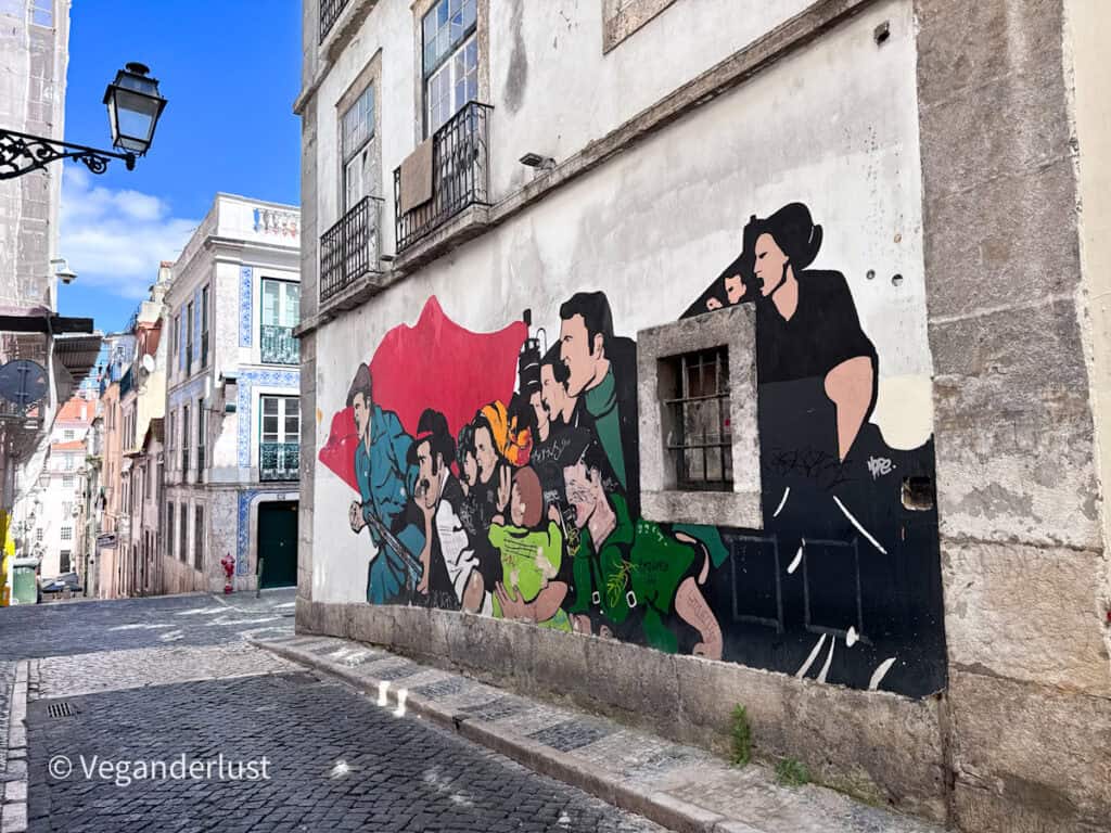 Street mural of a group of illustrated people in bold colors walking along a cobblestone Lisbon street lined with historic buildings. A small window interrupts the artwork, blending into the scene.