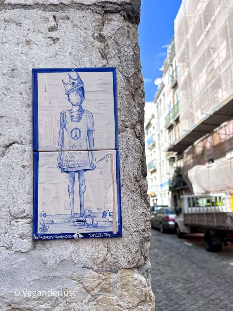 Blue tile style street art shows a figure wearing a crown and peace symbol shirt with handwritten text beneath. The artwork is mounted on a stone wall along a narrow street.