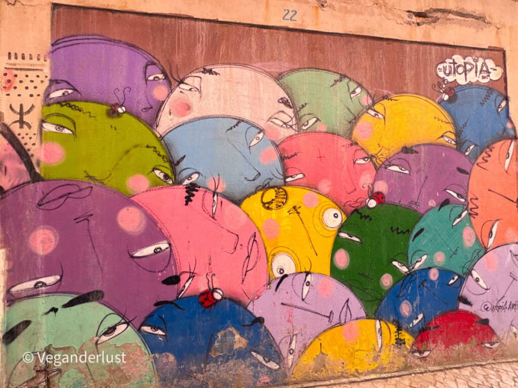 Colorful mural of overlapping abstract faces in pink, green, yellow, and blue tones with the word "utopia" written in the corner. The faces fill the entire wall in a playful layered composition.