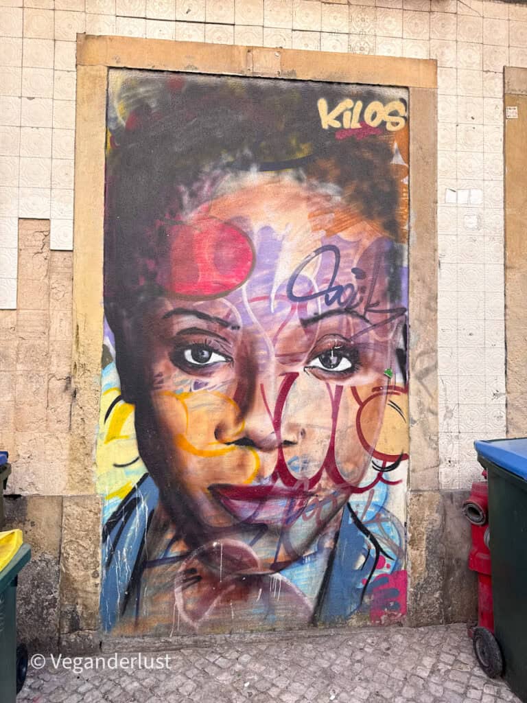Portrait style graffiti of a woman's face layered with colorful tags and paint strokes on a worn wall. Nearby trash bins and textured tiles add contrast to the vibrant artwork.