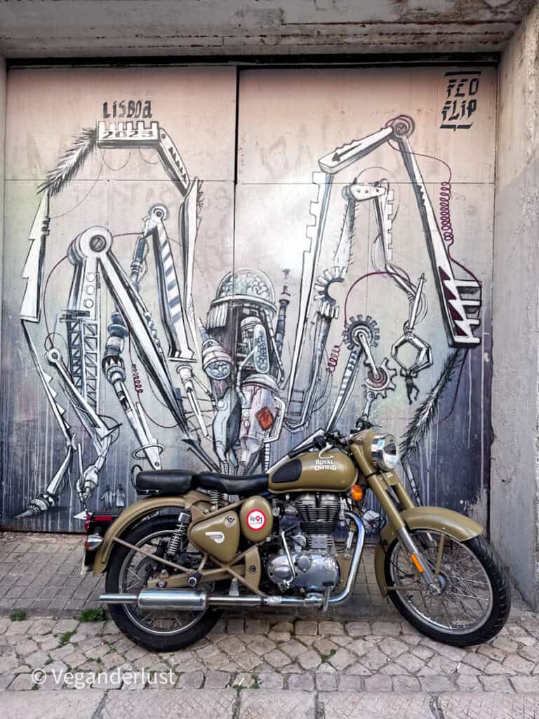 Vintage motorcycle parked in front of a detailed mural of mechanical arms and industrial shapes forming a futuristic figure. The contrast between real bike and painted machinery creates a striking urban scene.