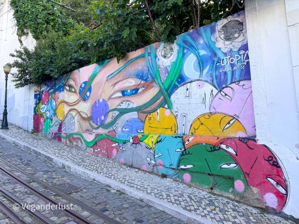 Long mural along a tram track wall featuring stylized faces and flowing shapes in bright colors like blue, green, and pink. Trees above the wall and cobblestone path frame the street art scene.