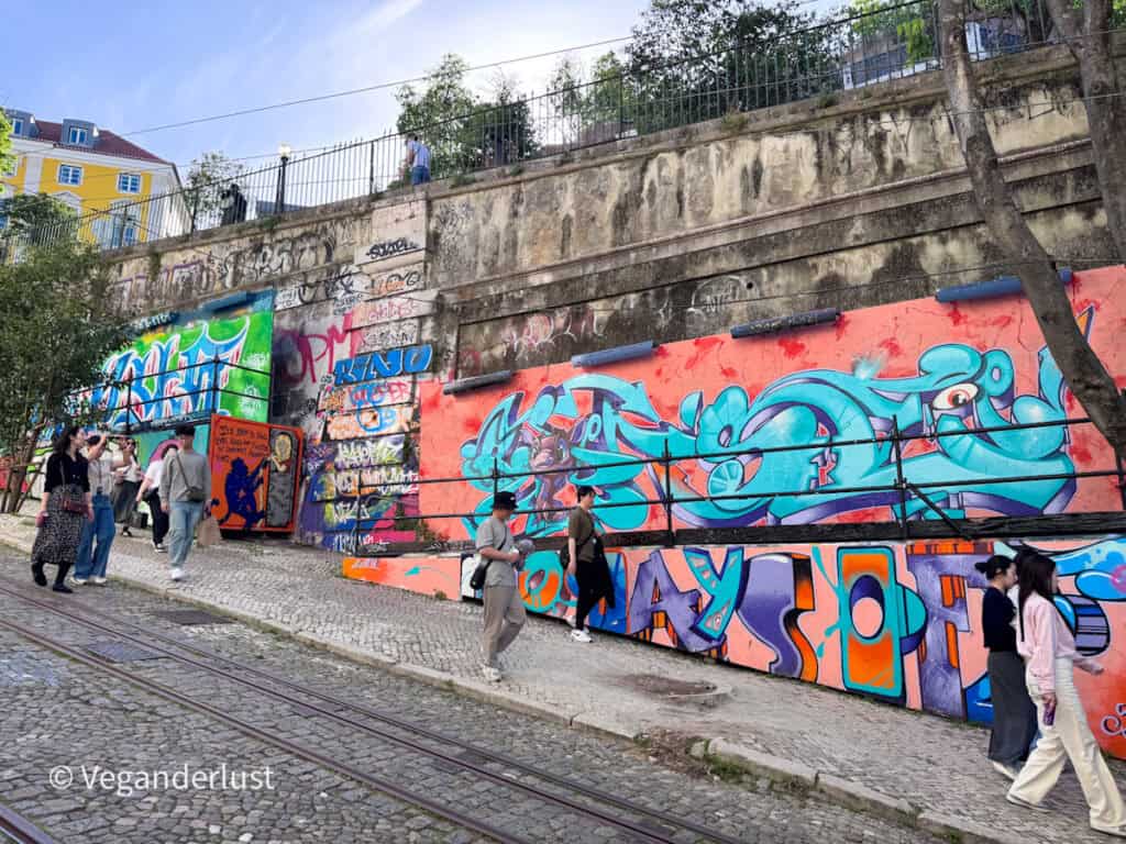 Graffiti covered wall beside tram tracks with people walking past colorful tags and large lettering. The urban setting shows Lisbon street art integrated into everyday city life.