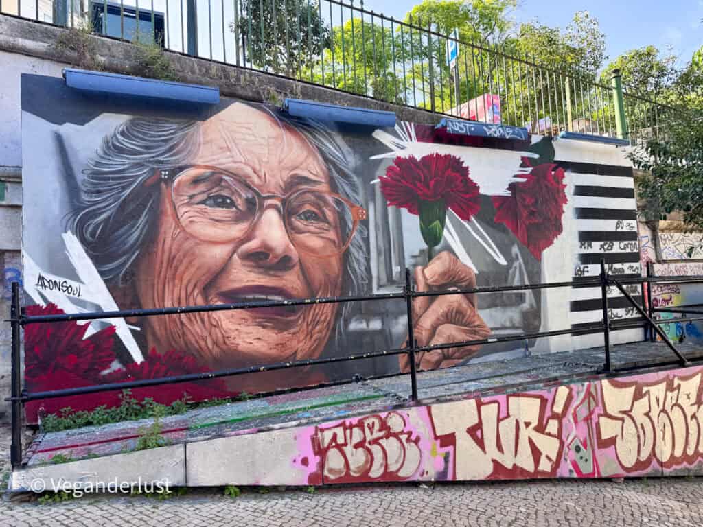 Realistic mural of an elderly woman wearing glasses holding red carnations, painted on a wall above layered graffiti tags. The detailed portrait contrasts with the rough urban textures and railings in this Lisbon street art scene.