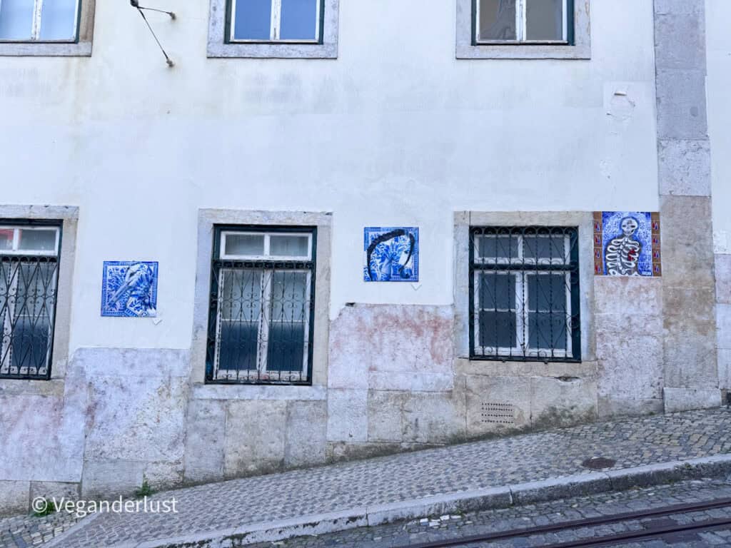 White building facade with barred windows decorated by small blue tile artworks featuring birds and a skeleton. The cobblestone street and tram tracks emphasize Lisbon’s blend of traditional tiles and modern street art.
