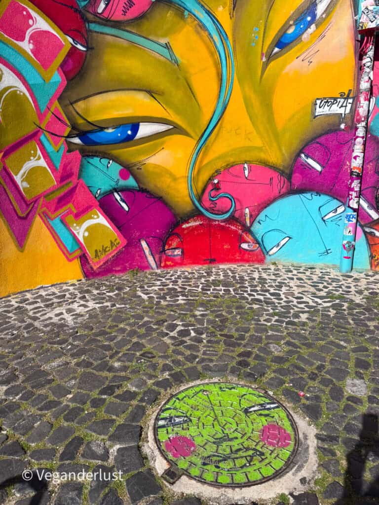 Close up of a vibrant mural with stylized faces in bright pink, yellow, and blue surrounding a larger pair of eyes. A cobblestone ground and painted manhole cover appear in the foreground.