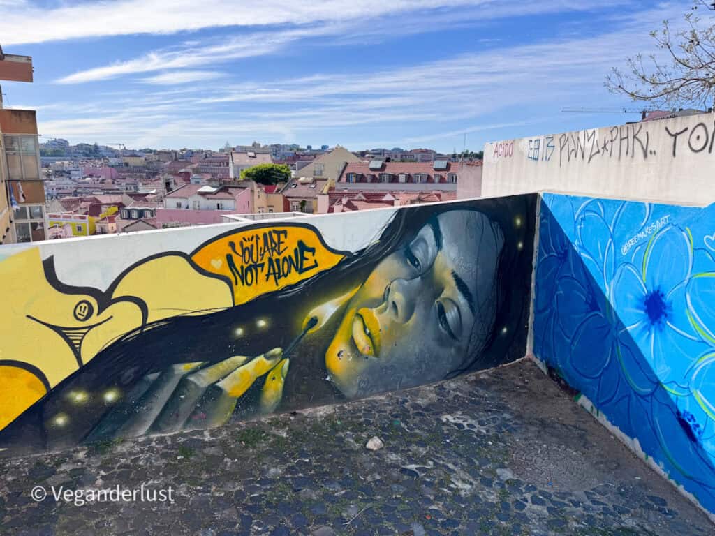 Mural of a woman lighting a match with glowing yellow tones and the text "YOU ARE NOT ALONE" written beside her. The artwork overlooks a scenic view of Lisbon rooftops in the background.