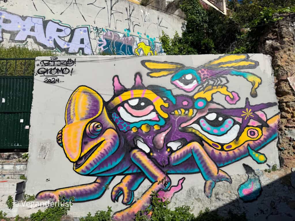 Abstract graffiti mural of a colorful creature with multiple eyes and limbs in purple, yellow, and teal tones. The design fills a white wall with layered tags and textures around it.