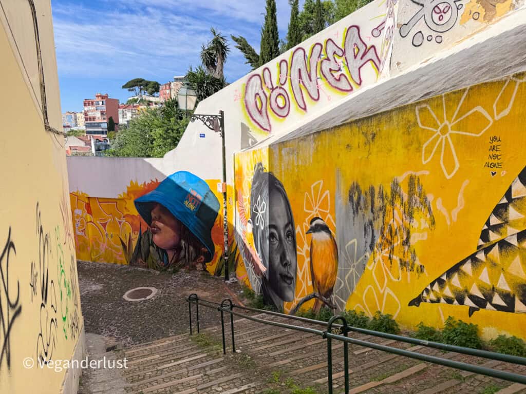 Sloped street lined with murals including a woman in a blue bucket hat and a black and white portrait beside a painted bird. The bright yellow wall and layered artwork create a vibrant Lisbon street art corner.