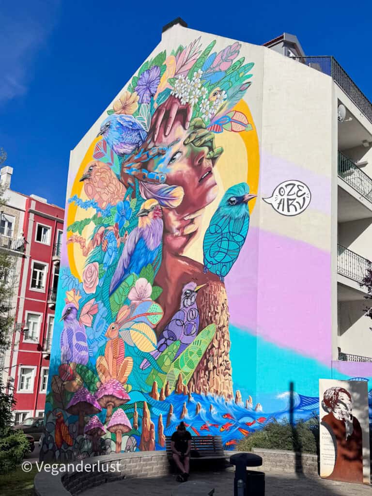 Large colorful mural of a woman surrounded by birds, flowers, and ocean elements painted on the side of a building. The vibrant artwork transforms the Lisbon street corner into an immersive street art landmark.