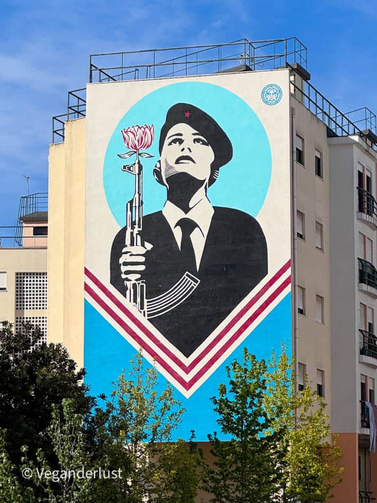 Bold graphic mural of a woman holding a rifle with a flower at the barrel set against a blue circular background. The striking political style artwork covers the full height of a Lisbon apartment building.