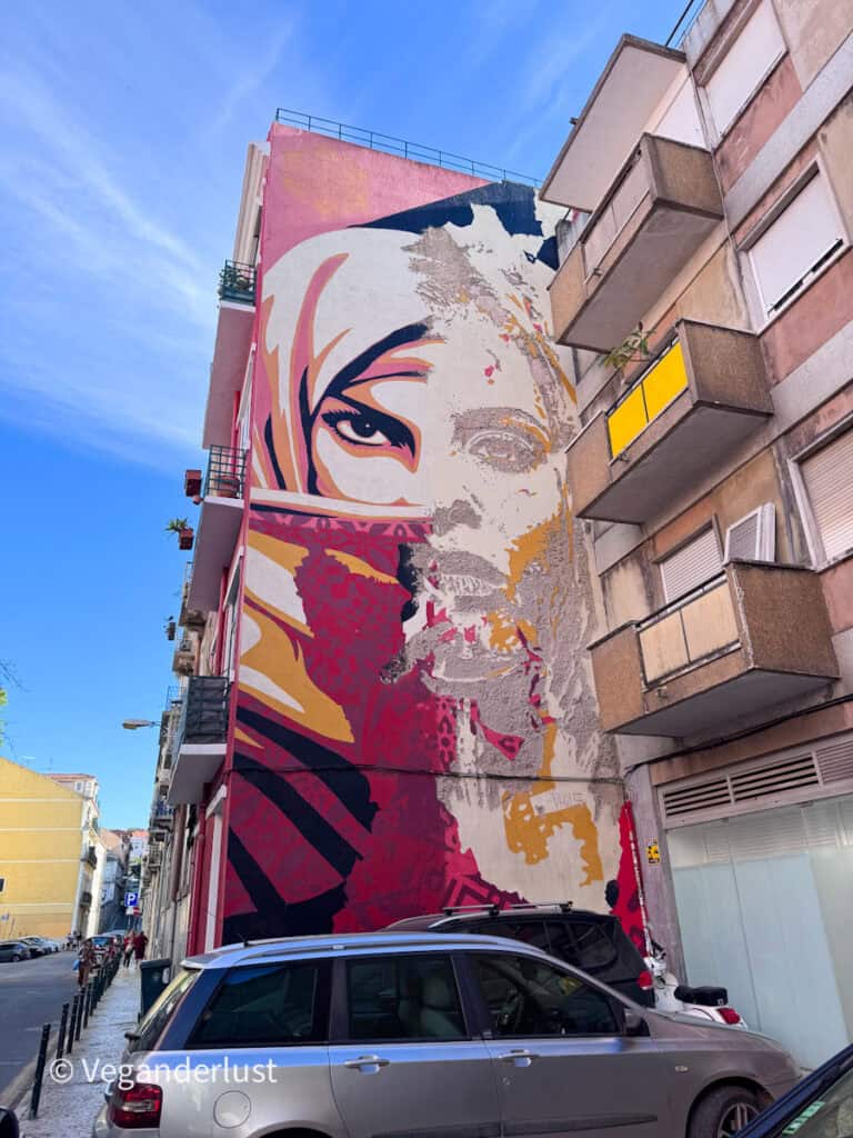 Weathered mural of a woman’s face in red, pink, and beige tones peeling away on the side of a tall building. Parked cars and balconies frame this aged Lisbon street art piece.
