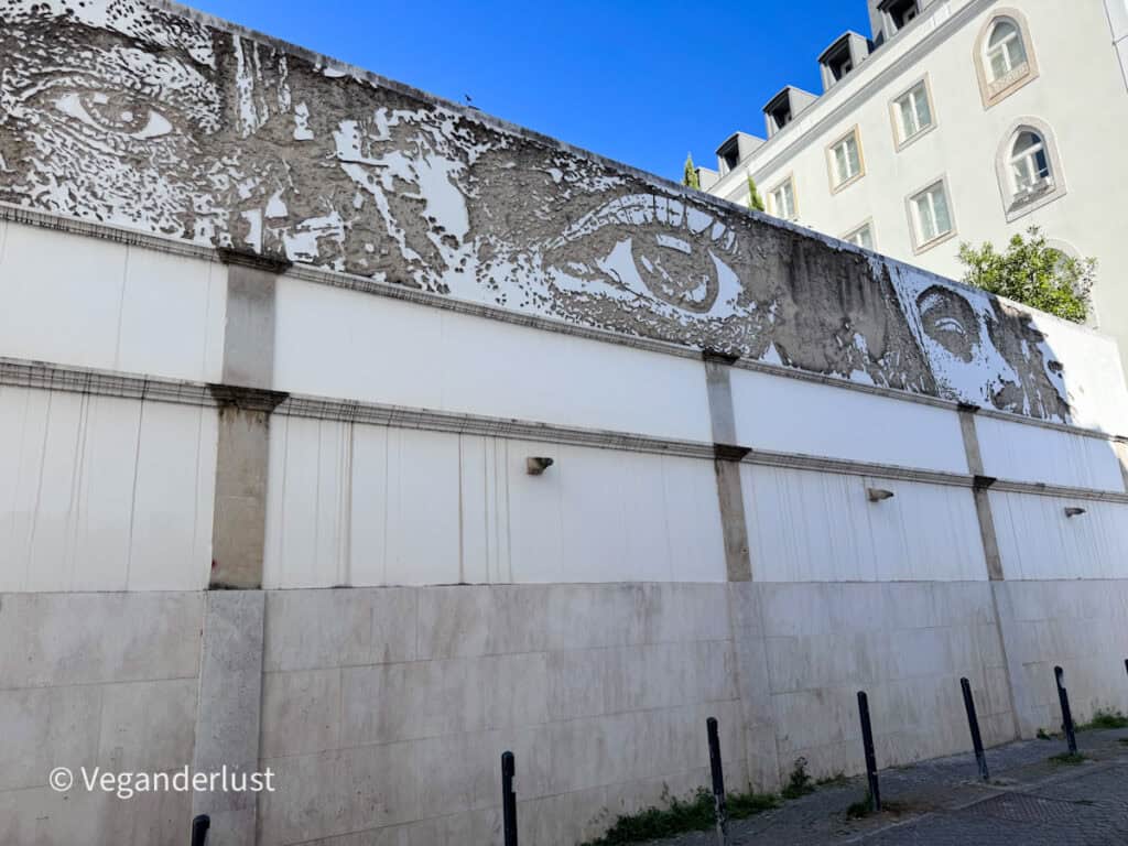 Large faded mural of a pair of eyes stretching across the top of a long wall with peeling paint textures. The subtle artwork blends into the architecture along a quiet Lisbon street.
