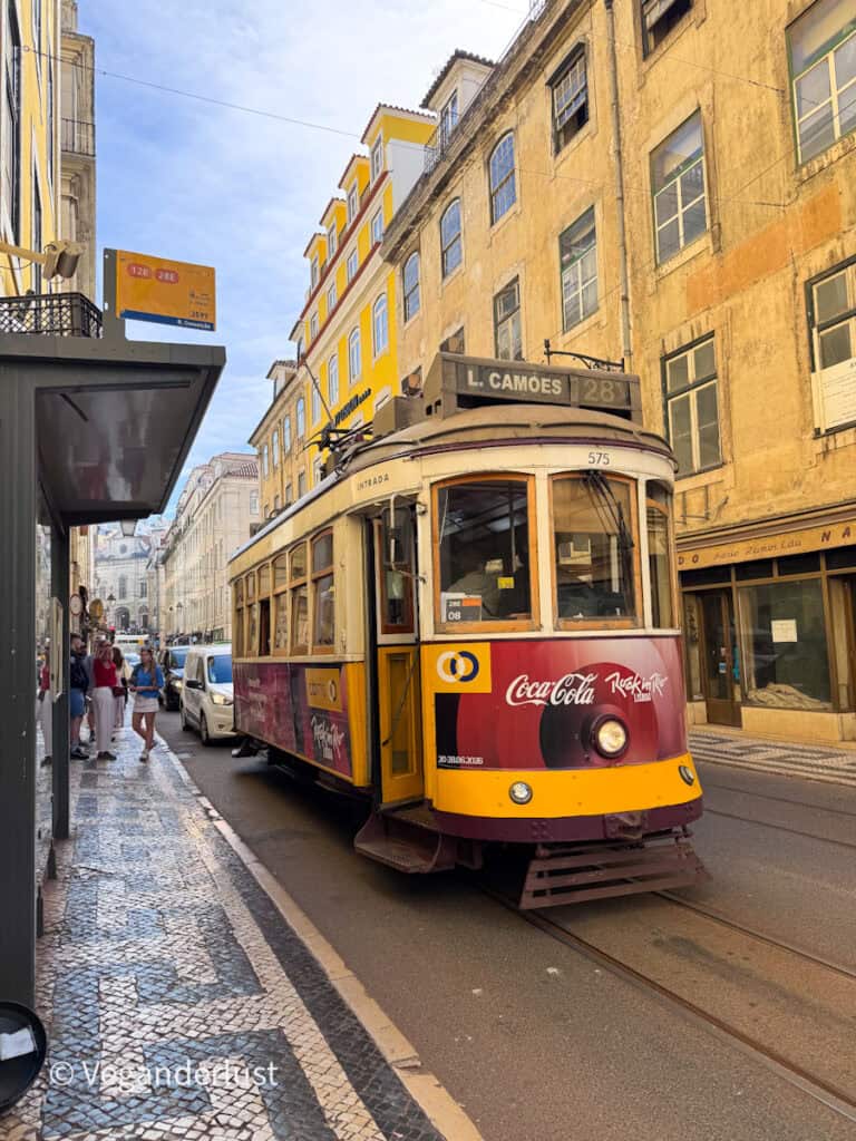 Classic yellow Lisbon tram labeled "L. Camões 28E" travels down a narrow street lined with pastel buildings and patterned tile sidewalks. People wait at a stop beside the tram while cars follow behind on the tracks.