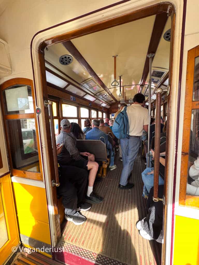 Interior of a Lisbon tram with passengers seated on wooden benches and others standing in the aisle holding overhead straps. Sunlight streams through the windows, highlighting the narrow vintage carriage and crowded atmosphere.