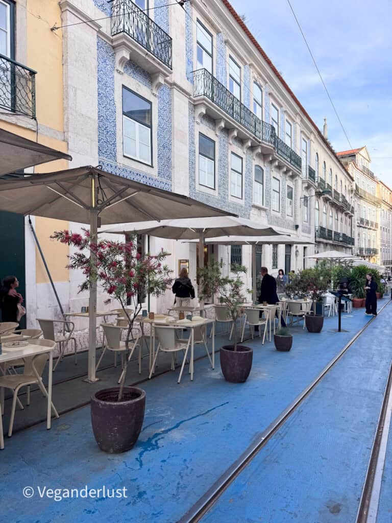 Outdoor restaurant seating with beige chairs and large umbrellas lines a blue-painted street beside a building covered in blue and white tile patterns with wrought iron balconies. A few people walk past while potted plants are spaced along the edge of the dining area.