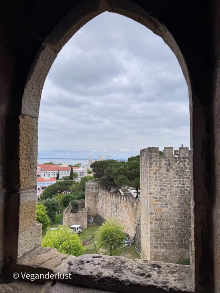 View through a stone archway frames castle walls and towers surrounded by greenery, with Lisbon buildings and the river visible in the distance. The cloudy sky adds a muted tone to the historic scene.