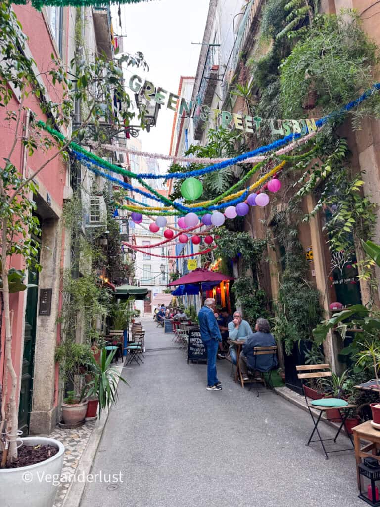 Narrow alley filled with greenery and small café tables where people sit and chat, decorated with hanging lanterns and garlands overhead. A sign strung across the alley reads "GREEN STREET LISBON" above the lively scene.