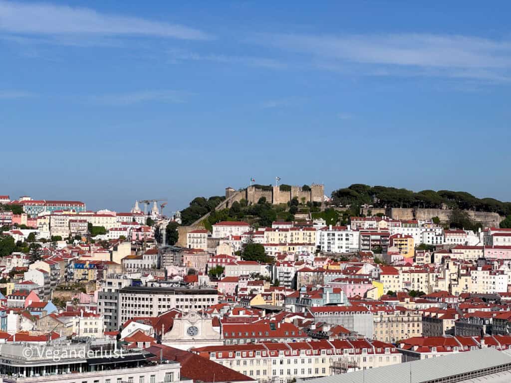 Close view of Lisbon’s colorful buildings with red tiled roofs clustered together beneath a hilltop castle surrounded by trees. The bright sky highlights the layered architecture and historic skyline.