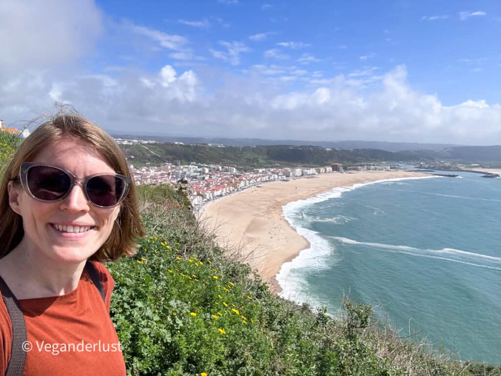 Person wearing sunglasses smiling at the camera while standing on a grassy cliff overlooking a wide beach and coastal town. Waves curve along the shoreline and clusters of buildings stretch along the bay in the background.
