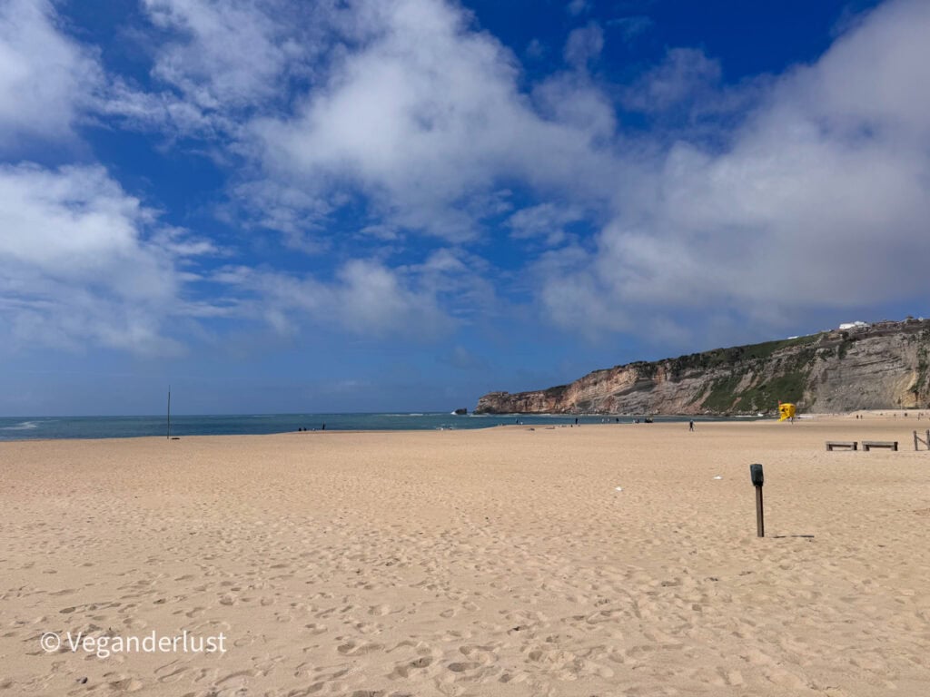 Wide sandy beach stretching toward the ocean under a blue sky with scattered clouds. Cliffs rise along the right side and a yellow lifeguard stand sits near a few distant people walking along the shore.