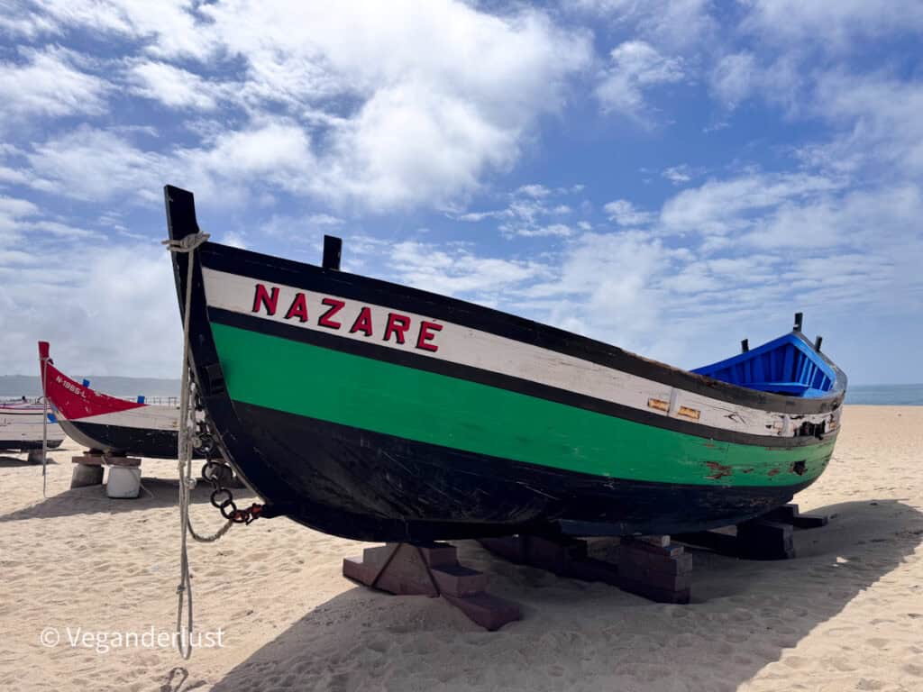 Traditional wooden fishing boat painted black green and white resting on the sand. The word "NAZARE" is painted in red letters on the side with ropes and another boat visible behind it.