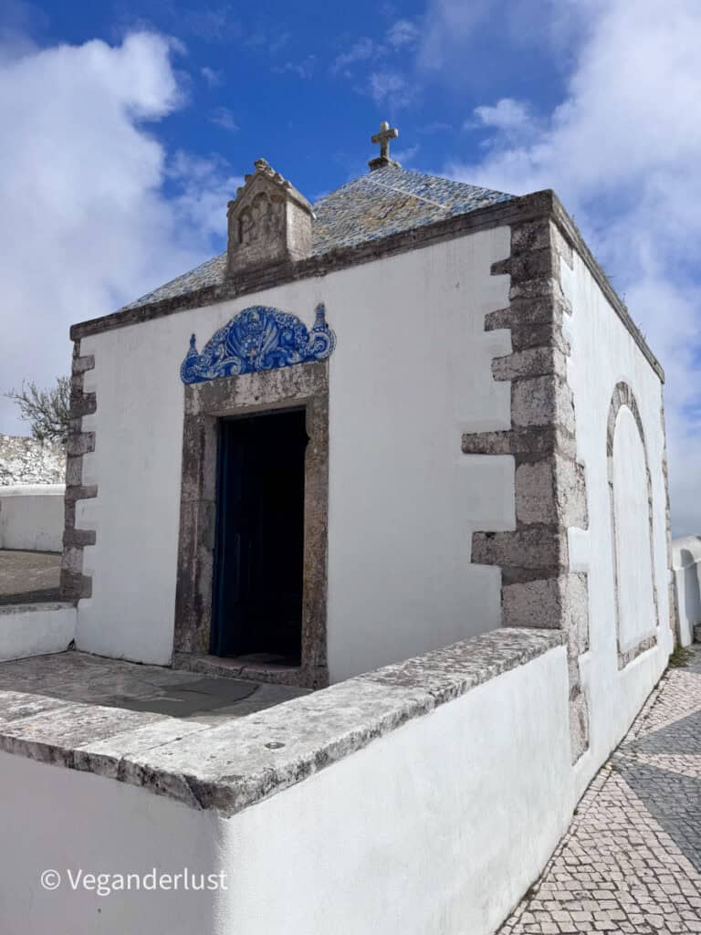 Small white chapel with stone corners and a cross on the roof viewed from the entrance. Blue decorative tiles frame the doorway and a narrow stone walkway runs along the side.
