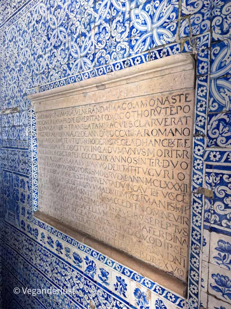 Close up of a stone plaque set into a wall covered in blue patterned tiles. The plaque features engraved text in multiple lines surrounded by intricate floral tile designs.
