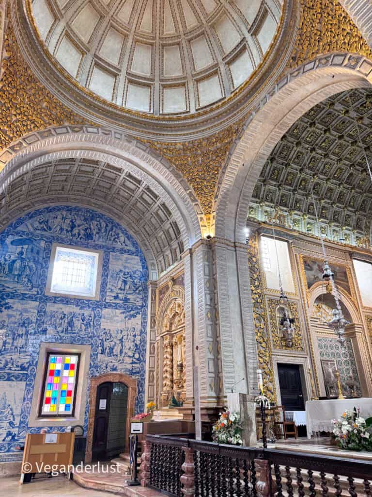Ornate church interior with high arched ceilings decorated in gold details and blue tile murals. A colorful stained glass window and altar with flowers and chandeliers are visible.