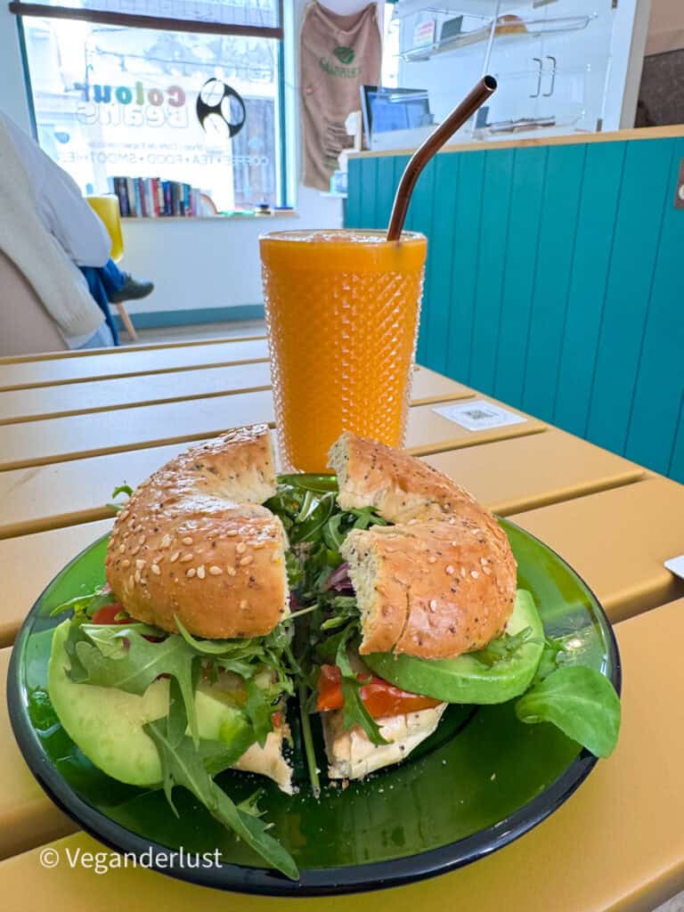 Bagel sandwich filled with greens and vegetables on a green plate beside a textured glass of orange juice with a metal straw. The meal sits on a wooden table inside a cafe with a window and seating in the background.