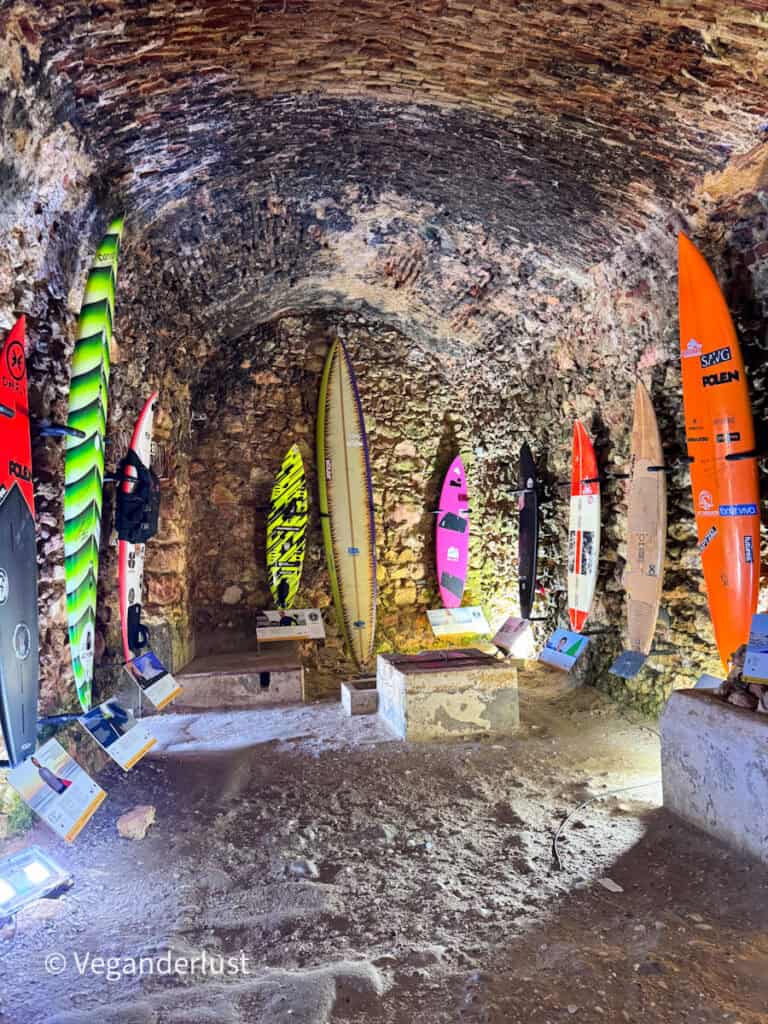 Stone chamber with a curved brick ceiling displaying a collection of surfboards propped upright along the walls. The boards vary in color and design with small informational plaques placed in front of each one.