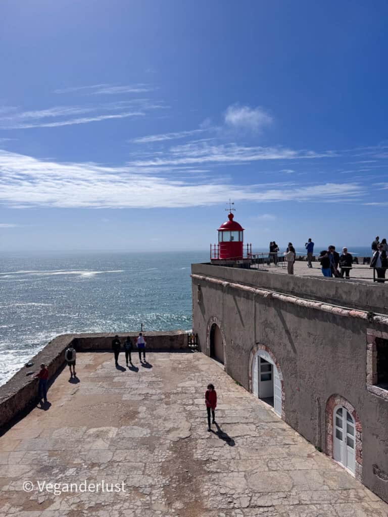 Red lighthouse perched on a stone fort overlooking the ocean with waves below. Visitors stand along the edge of the structure and walk across the open courtyard under a bright blue sky.