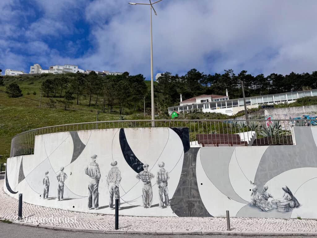 Curved outdoor wall painted with a monochrome mural of people standing and sitting in small groups. The artwork wraps around a street corner with a hillside of houses and trees rising behind it.
