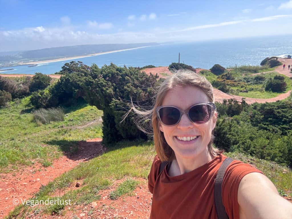 Person wearing sunglasses smiling for a selfie on a grassy cliff with red dirt paths. The ocean and a wide sandy coastline stretch out in the background under a bright sky.
