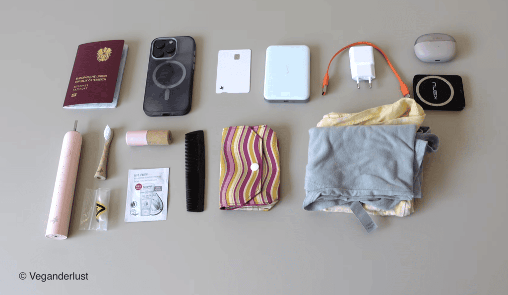 Travel essentials neatly arranged on a light surface including a passport, smartphone, credit card, power bank, charger with cable, wireless earbuds, tracking device, electric toothbrush, toothbrush, lip balm, comb, menstrual pad, small toiletry sample, and folded clothing. The layout shows a minimalist packing setup for daily use or travel with each item spaced evenly for easy viewing.