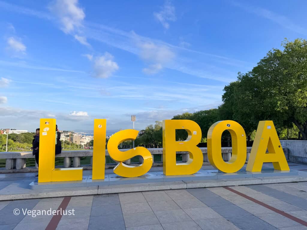 Large yellow letters spelling "LISBOA" stand on a terrace overlooking a park and city skyline, with a few people standing nearby taking photos. The bright sign contrasts against a blue sky with light clouds.